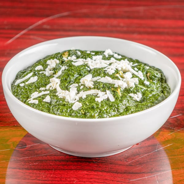 A white bowl of vibrant green Palak Paneer, topped with crumbled white paneer cheese, on a red wooden table.