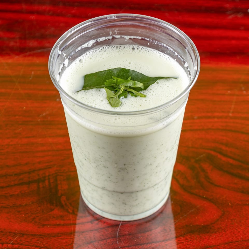 A plastic cup of Indian buttermilk, garnished with fresh green curry leaves, on a red wooden table.