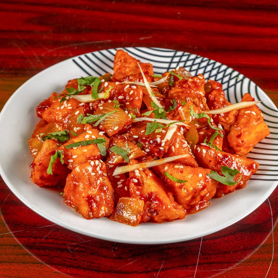 Indian chili paneer, a vibrant red dish with sesame seeds, cilantro, and ginger on a white plate.