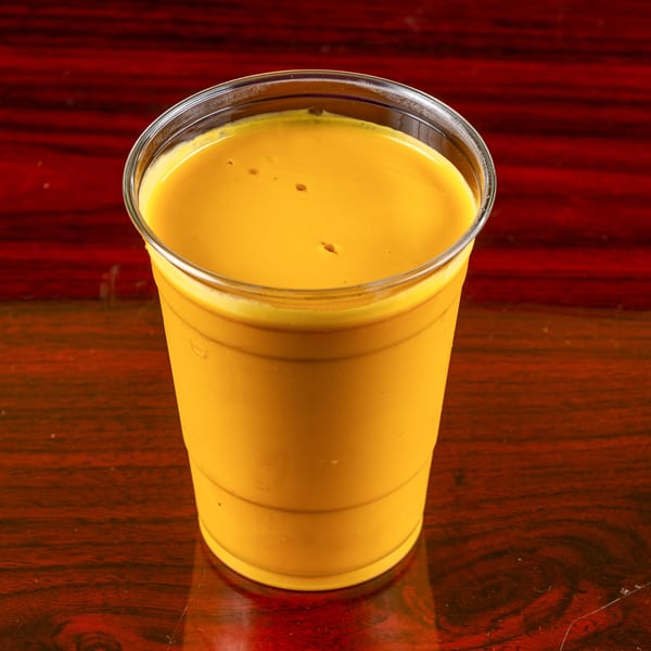A vibrant yellow mango lassi in a clear plastic cup sits on a dark wooden table.