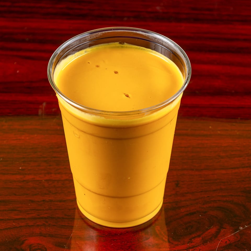 A vibrant yellow mango lassi in a clear plastic cup sits on a dark wooden table.
