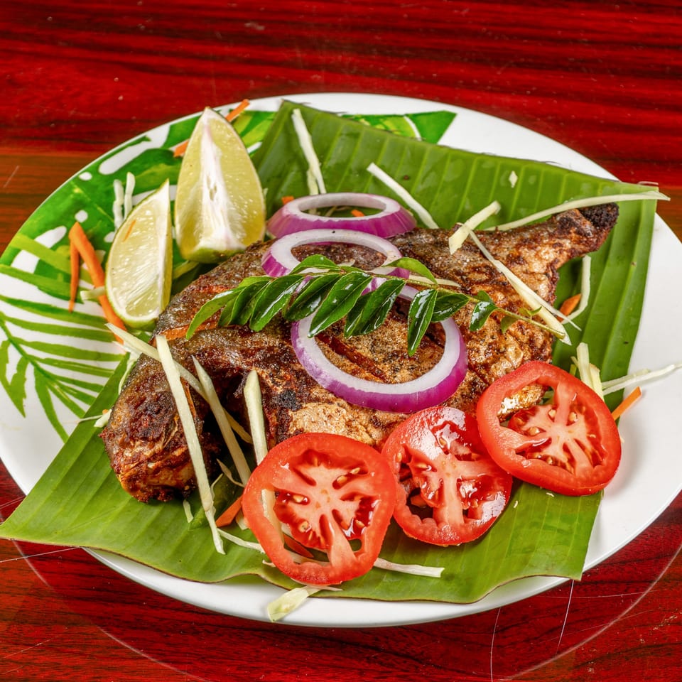 Indian fried fish on a banana leaf, garnished with lime, onions, tomatoes, and curry leaves.