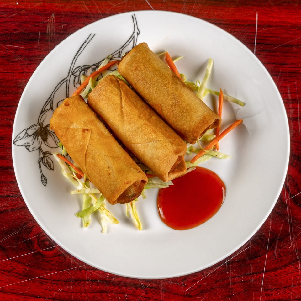 Three golden-brown spring rolls on a white plate with shredded cabbage, carrots, and a red dipping sauce.